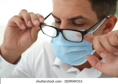 Man Wiping Foggy Glasses Caused By Wearing Medical Mask Indoors, Closeup