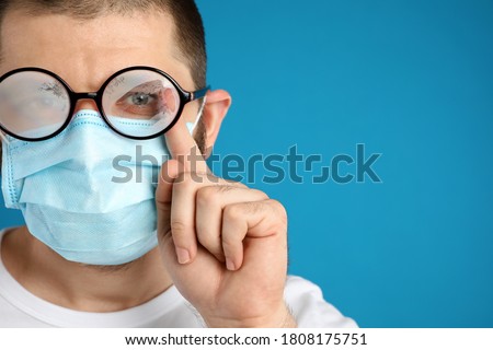 Man wiping foggy glasses caused by wearing disposable mask on blue background, space for text. Protective measure during coronavirus pandemic