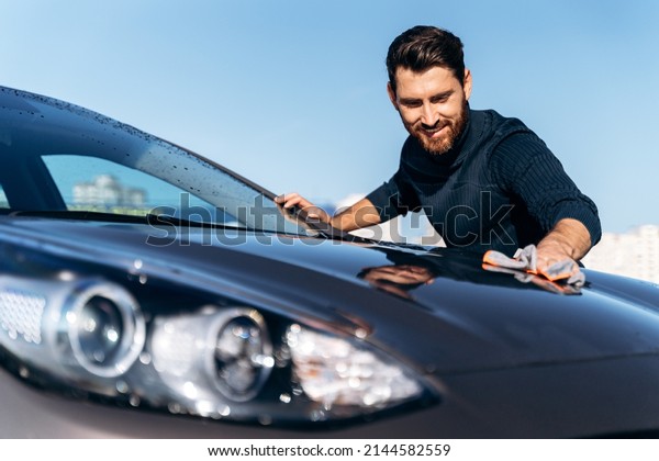 Man wiping his car at the street. Close up view of the\
handsome bearded man in casual wear washing car doors and hood with\
microfiber cloth. Car detailing wash during the sunny day concept\
