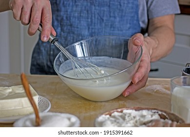 A Man Without A Face In An Apron Holds A Whisk In His Hands And Kneads Dough For Baking In The Kitchen. Authentic Home Cooking Hobby, Home Baker. Man Cooking In The Kitchen At Home