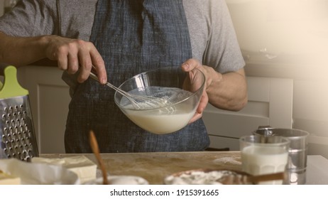 A Man Without A Face In An Apron Holds A Whisk In His Hands And Kneads Dough For Baking In The Kitchen. Authentic Home Cooking Hobby, Home Baker. Man Cooking In The Kitchen At Home