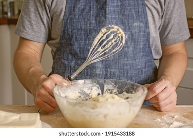 A Man Without A Face In An Apron Holds A Whisk In His Hands And Kneads Dough For Baking In The Kitchen. Authentic Home Cooking Hobby, Home Baker. Man Cooking In The Kitchen At Home