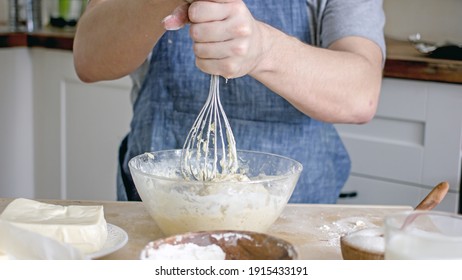 A Man Without A Face In An Apron Holds A Whisk In His Hands And Kneads Dough For Baking In The Kitchen. Authentic Home Cooking Hobby, Home Baker. Man Cooking In The Kitchen At Home