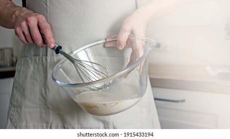 A Man Without A Face In An Apron Holds A Whisk In His Hands And Kneads Dough For Baking In The Kitchen. Authentic Home Cooking Hobby, Home Baker. Man Cooking In The Kitchen At Home