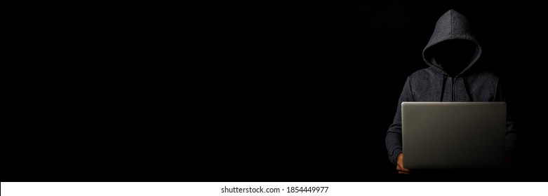 Man without a face in a hood holds a laptop on a dark background. Concept of cyber rogue, hacker. Banner - Powered by Shutterstock