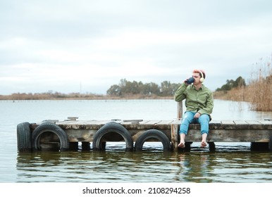 Man without shoes sits on a platform while drinking coffee and listening to music facing a lake