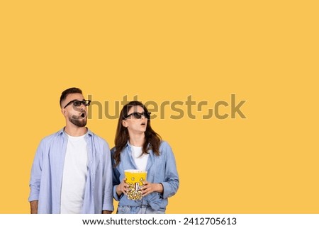 Man and woman in 3D glasses look up at free space in awe, holding popcorn bucket, suggesting an immersive movie experience, against vivid yellow background