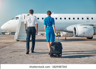 Man and woman in aviation uniform are walking to plane stairway with luggage before departure
