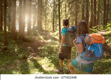 Man And Woman With Backpack Walking On Hiking Trail Path In Forest Woods During Sunny Day.Group Of Friends People Summer Adventure Journey In Mountain Nature Outdoors.Travel Exploring Alps,Dolomites