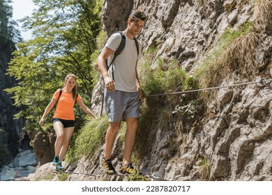 Man and woman with backpacks hiking carefully on the narrow, steep path above the rocky mountain river. Togetherness and outdoors pastime activity concept.