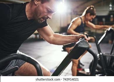 Man And Woman Behind Riding Cycling Machine In Hard Efforts Grimacing In Light Spacious Gym. 