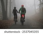 A man and a woman in bicycle helmets ride sports bicycles with their headlights on along the alley of an autumn park in cold foggy weather