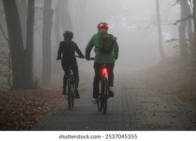 A man and a woman in bicycle helmets ride sports bicycles with their headlights on along the alley of an autumn park in cold foggy weather - Powered by Shutterstock