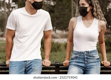 A man and woman, both wearing masks, sit on a bench outdoors. The man and woman, both in casual attire, engage in conversation. The man and woman enjoy a sunny day wearing white shirts for mockup. - Powered by Shutterstock