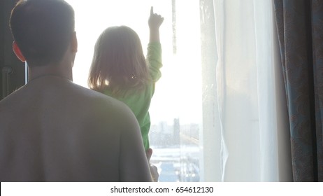 A Man, A Woman And A Child Look Out The Window And Play With The Rays Of The Sun. Love In The Family. Father, Mother And Their Child