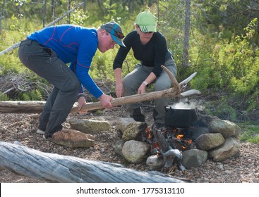 A Man And A Woman Cook Fish Soup In Nature. The Pot Is Carefully Removed From The Fire. Picnic In Nature. Camping Life And Kitchen.