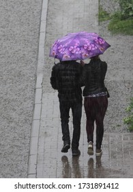 A man and woman couple in black jackets under one bright pink umbrella walking together along the sidewalk near a large puddle on a cold summer rainy day