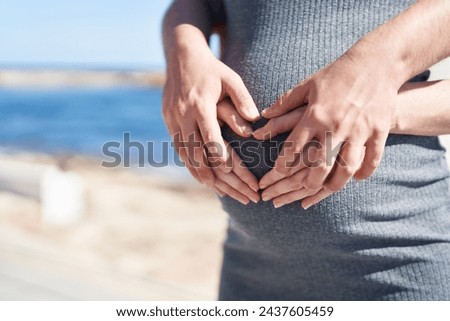 Man and woman couple hugging each other doing heart gesture on belly at seaside