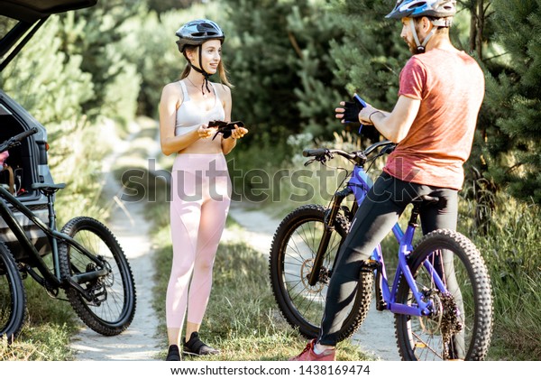 Man and woman cyclists wearing gloves and helmet,\
preparing for the bicycle riding while standing on the forest road\
during the summer time