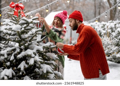 Man and woman decorate Christmas tree with festive balls, while preparing for a winter holidays at snowy backyard of their house. Happy family celebrating New Year's holidays - Powered by Shutterstock