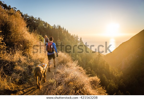 Man, woman, and dog hike in Big Sur, CA as the sun
sets over the ocean.