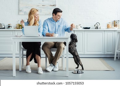 Man And Woman Drinking Coffee And Playing With Their Dog By Kitchen Table With Laptop