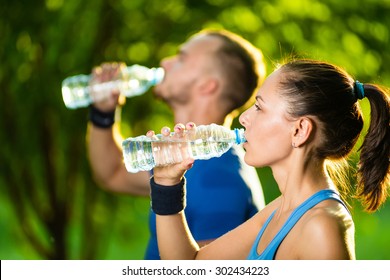 Man And Woman Drinking Water From Bottle After Fitness Sport Exercise. Smiling Couple With Bottles Of Cold Drink Outdoors