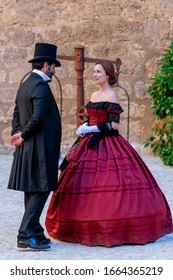 A man and a woman, elegantly dressed, in 19th-century clothes, walk and talk, in the courtyard of a palace
