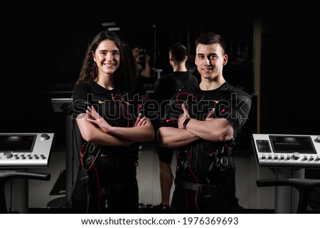 man and a woman in an EMS vest are standing side by side and smiling. 2 trainers in gym.