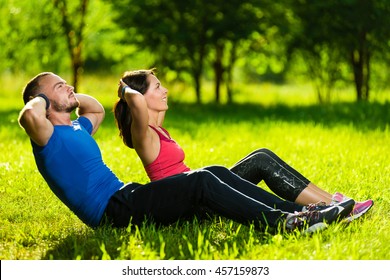 Man And Woman Exercising At The City Park. Beautiful Young Multiracial Couple. Sit Ups Fitness Couple Exercising Outside In Grass. Fit Happy People Working Out Outdoor.