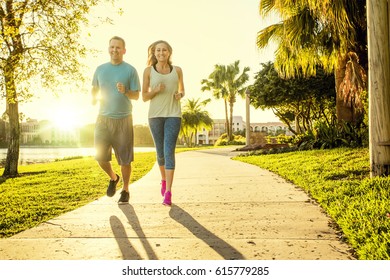 Man And Woman Exercising And Jogging Together At The Park. Happy And Smiling As They Run Along The Path During Sunset On A Warm Summer Day