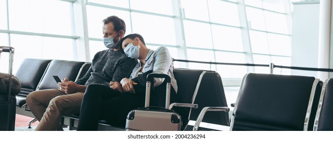 Man And Woman With Face Masks Sitting At Airport Terminal Waiting Lounge. Traveler Couple Stranded During Corona Virus Outbreak At Airport.