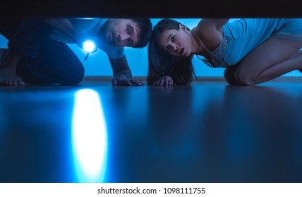 The Man And A Woman With A Flashlight Looking Under The Bed. Evening Night Time