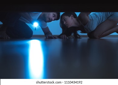 The Man And A Woman With A Flashlight Looking Under The Bed. Evening Night Time