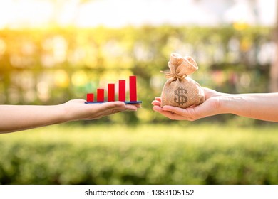 Man And Woman Hand Hold A Money Bag And Red Bar Graph With Growing Value Filed Together In The Public Park, Currency And Financial Business Investment To Future And Fund Concept.