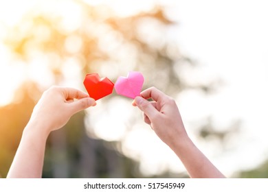 A man and woman hand holding a red heart and pink heart with love in sunlight in the public park.