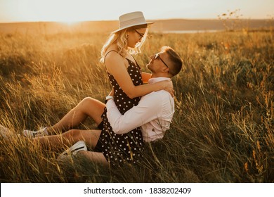 A Man With A Woman In A Hat Hug And Kiss In The Tall Grass In The Meadow. A Couple Of Fair-haired Fair-skinned People In Love Are Resting In Nature In A Field At Sunset.