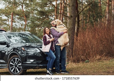 Man With Woman Have Fun With Their Dog Outdoors In Forest Near Modern Black Car.