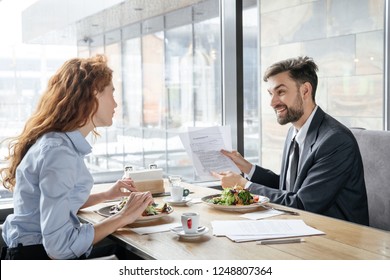 Man and woman having business lunch at restaurant sitting at table near window eating fresh salad drinking coffee discussing project smiling cheerful