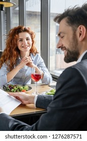Man and woman having business lunch at restaurant sitting at table eating vegetable salad drinking red wine businessman close-up reading contract smiling while businesswoman looking camera happy.