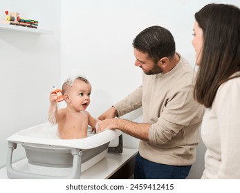 A man and a woman are helping a baby bathe. The baby is in a bathtub and the man is holding a toy - Powered by Shutterstock