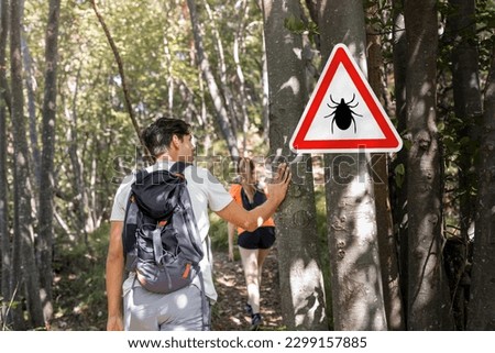 Man and woman hiking in Infected ticks forest with warning sign. Risk of tick-borne and lyme disease.