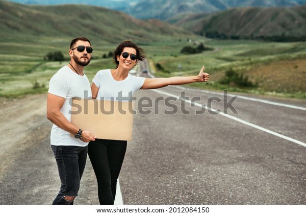 A man and a woman are hitchhiking. Wanderlust,\
autostop journey concept. Young people traveling hitchhiking.\
Hitchhiking couple on the road. Hitchhiking trip. Car travel. Two\
by the road. Copy space