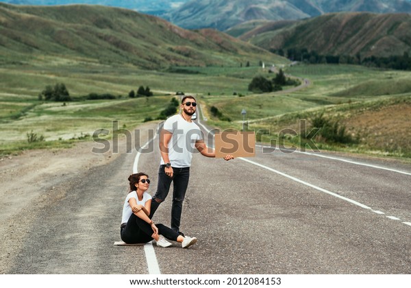 A man and a woman are hitchhiking. Wanderlust,\
autostop journey concept. Young people traveling hitchhiking.\
Hitchhiking couple on the road. Hitchhiking trip. Car travel. Two\
by the road. Copy space
