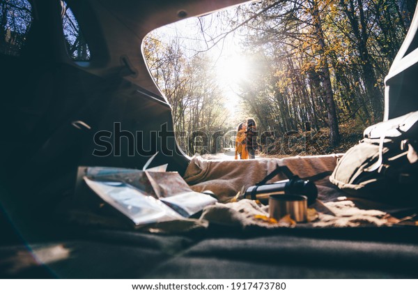 Man and woman hugging along empty rural road among\
autumn trees in sunny forest view through open rear door of modern\
car.