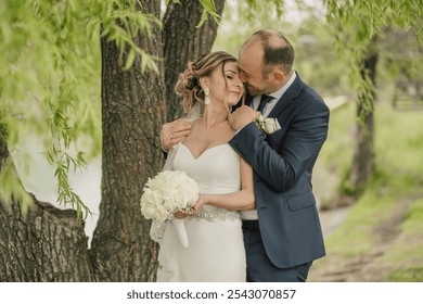 A man and woman are hugging under a tree. The man is wearing a blue suit and the woman is wearing a white dress - Powered by Shutterstock