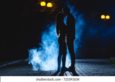 The Man And Woman Kissing On The Street On A Blue Smoke Background. Night Time
