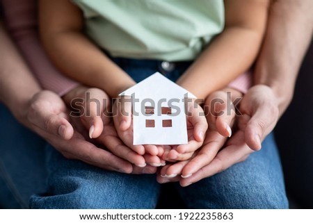 Man, woman and little child holding cutout paper house figure in hands, conceptual image for family housing, home mortgage, real estate, insurance or adoption, closeup shot