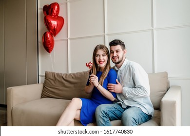 Man And Woman With Lollypop Candy At Home On Sofa With Heart Shaped Balloons