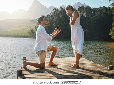 Man, woman and marriage proposal by lake on vacation with surprise, wow or happiness in sunshine. Couple, engagement and offer ring in nature for romance, love and happy on holiday in summer by water